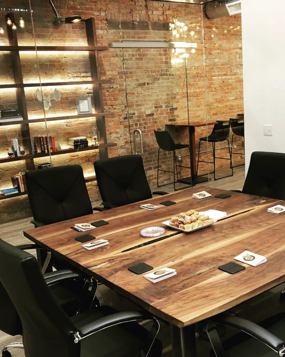 The conference room at The Lodge set for a meeting, with a live-edge walnut table, leather chairs, and the warm backlit bookshelf wall behind